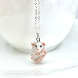 May include: A white ceramic mouse pendant necklace with a pink tail, hanging from a silver chain.
