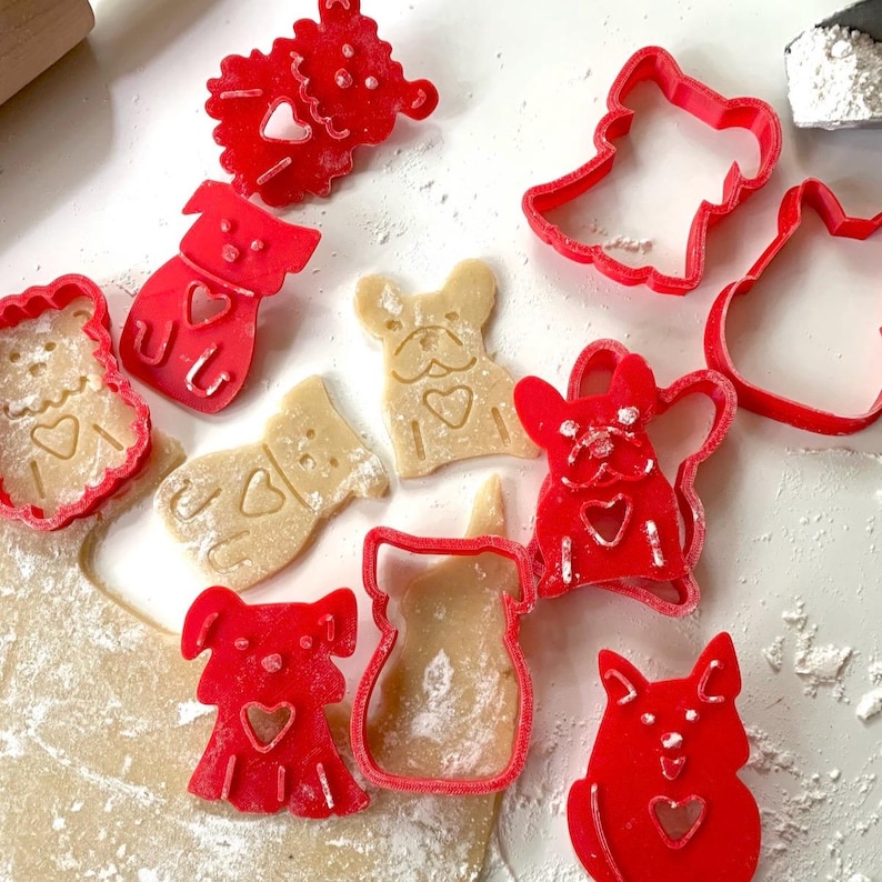 May include: Red plastic cookie cutters in the shapes of dogs, a heart, and a snowman. The cutters are on a white surface with a few cookies that have been cut out using the cutters.
