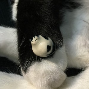 May include: A small, rounded ceramic figurine shaped like a curled-up cat with a smooth, glossy finish. The figurine is white with distinct black patches and spots, featuring painted facial details including closed eyes and small ears.