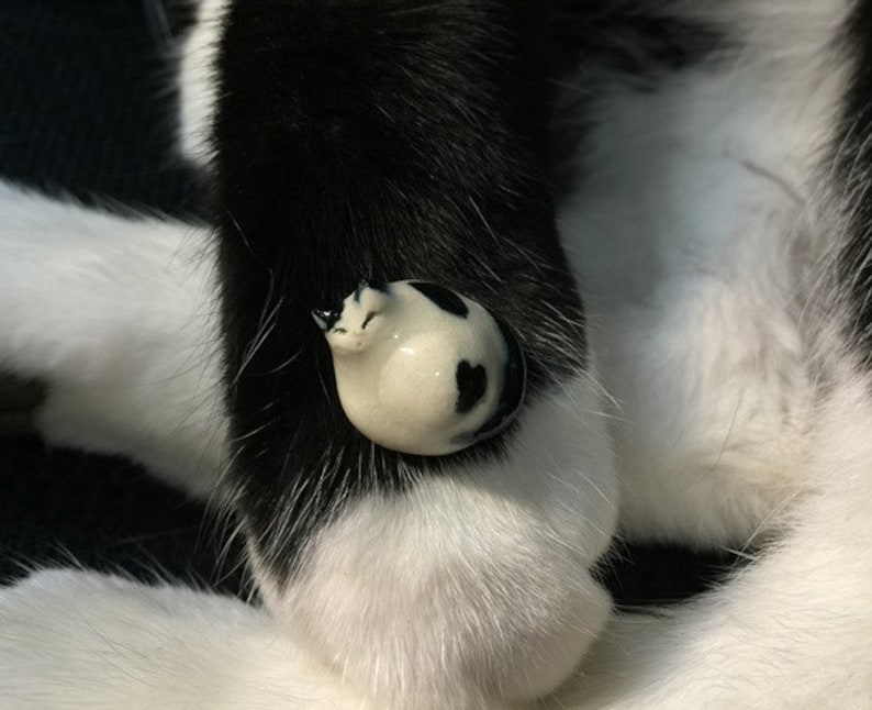 May include: A small, rounded ceramic figurine shaped like a curled-up cat with a smooth, glossy finish. The figurine is white with distinct black patches and spots, featuring painted facial details including closed eyes and small ears.