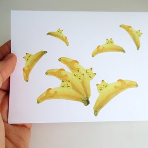May include: A white card with yellow watercolor illustrations of cats curled up inside bananas. The cats have black eyes and whiskers.