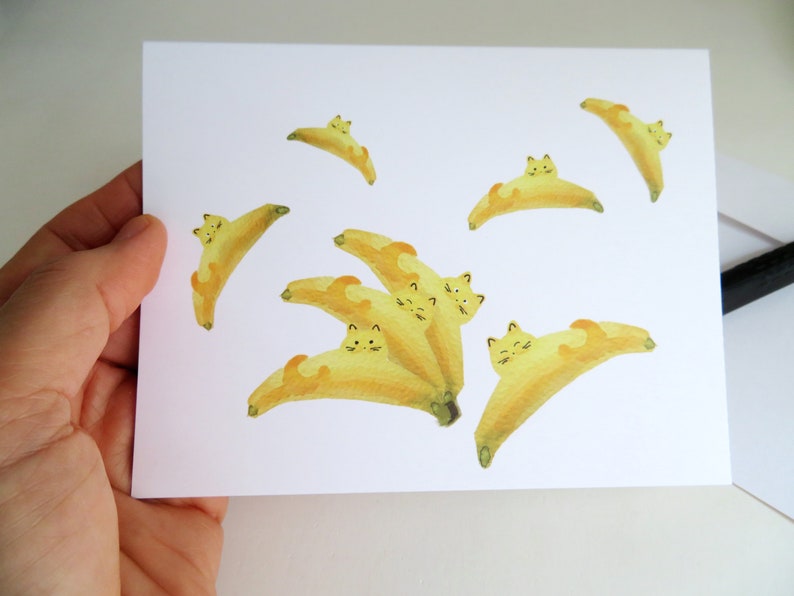 May include: A white card with yellow watercolor illustrations of cats curled up inside bananas. The cats have black eyes and whiskers.