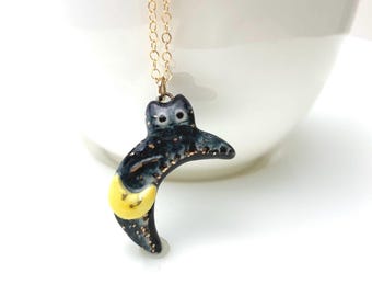 May include: A necklace featuring a black, crescent-shaped ceramic pendant with gold glitter accents, a small yellow crescent moon detail, and two white dots representing eyes, suspended from a thin gold-toned metal chain.