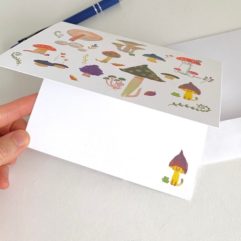 Op de afbeelding: Een witte kaart met een kleurrijke illustratie van paddenstoelen en kikkers. De kaart heeft een witte envelop. De illustratie toont paddenstoelen in verschillende kleuren, waaronder rood, oranje, paars en bruin. De paddenstoelen hebben witte stippen en zijn omringd door groene bladeren en ranken. Er zijn ook kleine kikkers in groen en geel.