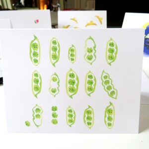 May include: A white card featuring a watercolour illustration of green pea pods with cute faces. The pea pods are arranged in a variety of positions and sizes.