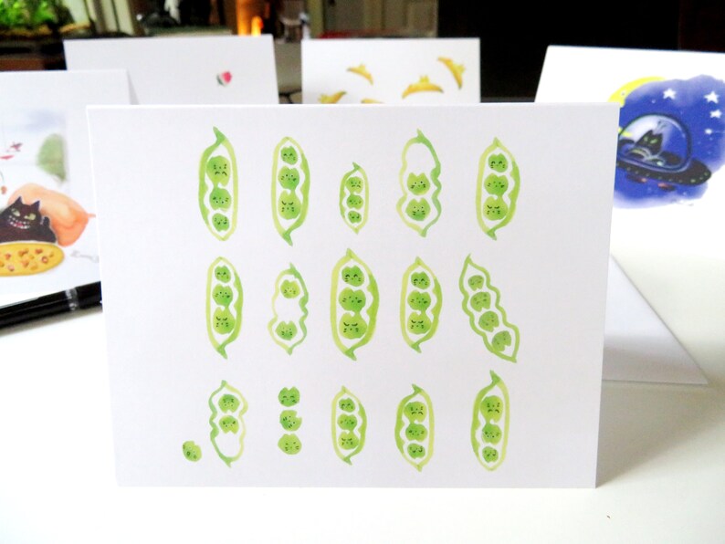 May include: A white card featuring a watercolour illustration of green pea pods with cute faces. The pea pods are arranged in a variety of positions and sizes.