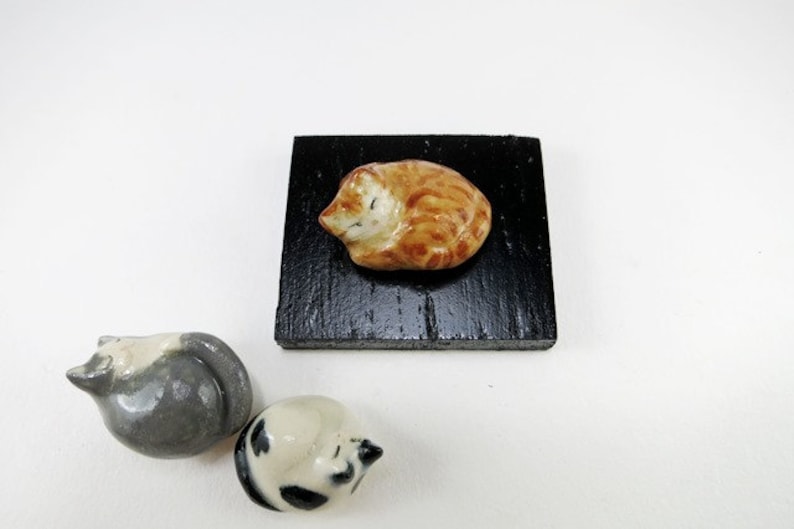 May include: A small orange ceramic cat figurine resting on a square black wooden base, accompanied by two additional ceramic cat figurines, one grey and white and one black and white, positioned separately on the surface.