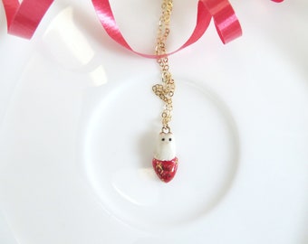 May include: A gold chain necklace with a white ceramic owl pendant. The owl is wearing a red heart-shaped dress with gold accents.