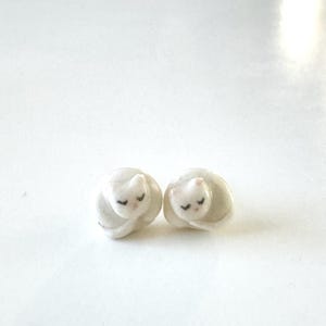 May include: Two small, white cat figurines with closed eyes and pink accents. The cats are curled up, appearing to be sleeping. The figurines are on a white surface.