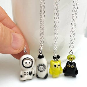 May include: Four whimsical pendant necklaces with silver chains. The pendants include a white astronaut cat, a white and black rocket ship cat, a yellow owl-like creature, and a black cat with a green eye. The necklaces are displayed against a white background.
