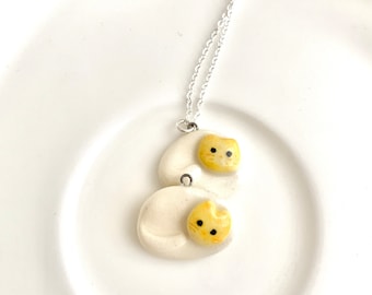 May include: A white ceramic necklace with a silver chain. The pendant is shaped like two cats, one on top of the other. The cats are yellow with black eyes and noses.