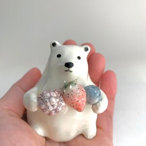 May include: A white ceramic polar bear figurine holding a strawberry, a blueberry, and a blackberry. The bear is sitting on a hand.