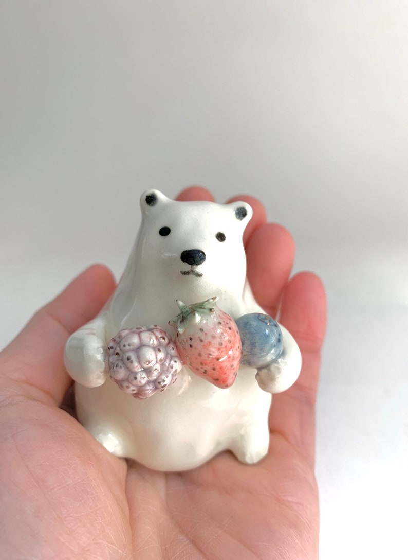 May include: A white ceramic polar bear figurine holding a strawberry, a blueberry, and a blackberry. The bear is sitting on a hand.
