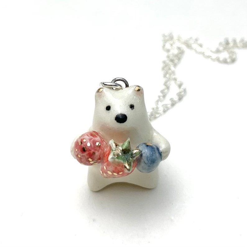 May include: White ceramic bear pendant necklace with a silver chain. The bear is holding a red and green strawberry and a blue blueberry.