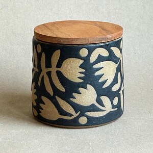 Pode incluir: Um pote cilíndrico de cerâmica com tampa de madeira. O pote é azul escuro com um padrão repetido de desenhos florais dourados. A tampa é de cor castanho claro.