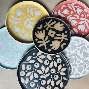 Puede incluir: Colección de platos de cerámica en varios colores, incluyendo amarillo, rojo, azul, negro y blanco. Cada plato presenta un diseño floral único en un color contrastante. Los platos son redondos y parecen hechos a mano.