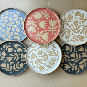 Puede incluir: Un juego de seis platos redondos de cerámica con diversos motivos botánicos en colores contrastantes. La colección incluye platos en azul, coral, blanco y gris oscuro, cada uno decorado con motivos florales o de bayas en beige claro.