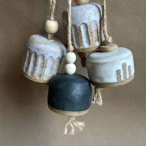 Puede incluir: Cuatro campanas de cerámica colgadas de una cuerda. Las campanas son en tonos de blanco, beige y negro, con un acabado rústico y texturizado. Cada campana está adornada con cuentas de madera. Las campanas cuelgan sobre un fondo neutro.