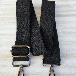 Puede incluir: Correa de bolso negra tejida con herrajes metálicos plateados. La correa presenta bucles metálicos rectangulares y cierres giratorios para la fijación. Fabricada con un material texturizado y duradero.