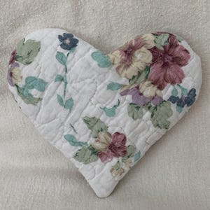 May include: A heart-shaped, quilted fabric item with a white background and a floral pattern in shades of pink, purple, green, and blue. The item has a quilted texture and is likely a decorative piece or a small mat.