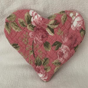 May include: A heart-shaped quilted item with a floral pattern in shades of pink, white, and green. The fabric features a rose design and is stitched with a quilted pattern. The item is likely a decorative piece or a small mat.
