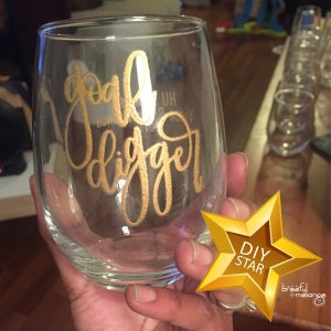 May include: A clear glass wine glass with the words "goal digger" written in gold lettering. The glass is being held by a hand. A gold star with the words "DIY STAR" is in the lower right corner of the image.