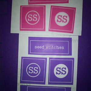 May include: Four labels with the text "seed stitches" and the initials "SS" in a circle. Two labels are pink with white text and initials, and two labels are purple with white text and initials.