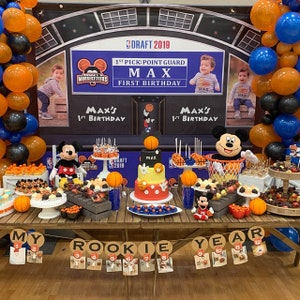 May include: A basketball-themed first birthday party table setting with a Mickey Mouse cake, a basketball hoop filled with candy, and a banner that reads "My Rookie Year".