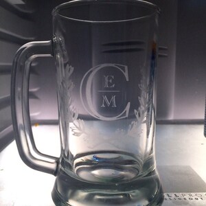 Puede incluir: Una jarra de cerveza de vidrio transparente con asa, con un dise&ntilde;o grabado de un monograma con las iniciales "E M" dentro de una "C" y rodeada de ramas con hojas.