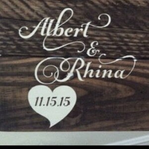 May include: A wooden sign with the names "Albert & Rhina" in white cursive lettering and a white heart with the date "11.15.15" in the center.