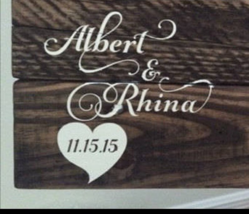 May include: A wooden sign with the names "Albert & Rhina" in white cursive lettering and a white heart with the date "11.15.15" in the center.