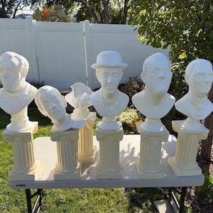 May include: Six white plaster busts of historical figures, each on a decorative column. The busts are displayed on a folding table outdoors, with a green lawn and white fence in the background. The busts are made of plaster.