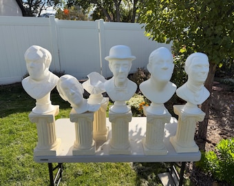 Puede incluir: Una colección de seis bustos esculpidos en blanco de varias figuras, cada uno montado sobre un pedestal estriado de color crema. Los bustos muestran diferentes rasgos faciales y peinados, incluida una figura que lleva un sombrero bombín.