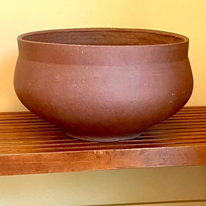May include: A large, round, brown ceramic planter sits on a wooden bench with a slatted top and rounded legs. The planter has a wide opening and a textured surface. The bench is made of wood and has a warm, reddish-brown tone.
