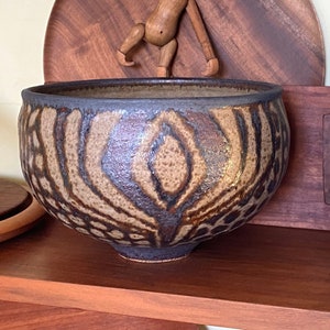 May include: A brown ceramic bowl with a geometric pattern sits on a wooden shelf. A wooden monkey figurine hangs from a wooden plate behind the bowl.