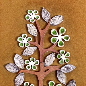May include: A brown felt wall hanging with a ceramic tree sculpture. The tree has white flowers with green centers and brown leaves.