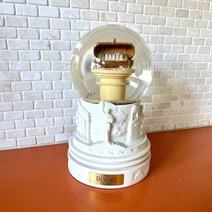 May include: A white snow globe with a gold-colored chest inside. The base of the snow globe is decorated with a white statue and the words "The Da Vinci Code".