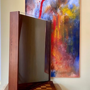 May include: A wooden frame with a dark glass panel inside. The frame is angled and rests on a wooden surface with a checkerboard pattern. A large abstract painting with red, yellow, blue, and brown colors hangs on the wall behind the frame.