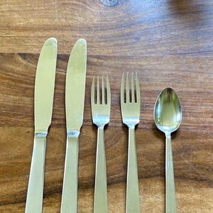 May include: A set of stainless steel flatware, including two knives, two forks, and a spoon, arranged on a wooden surface. The silverware has a simple, modern design with rectangular handles and a polished finish.