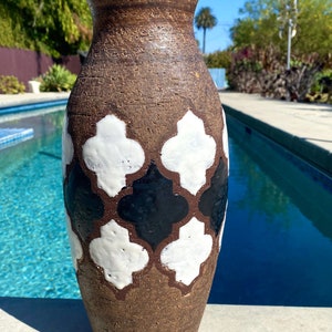 May include: A brown ceramic vase with a geometric pattern of white and black quatrefoils. The vase is sitting on a concrete surface near a swimming pool.