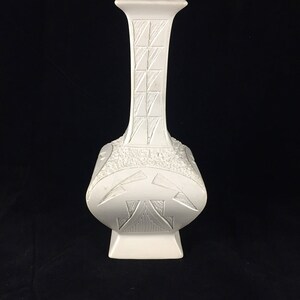 May include: A white ceramic vase with a geometric design. The vase has a square base and a tall, slender neck.