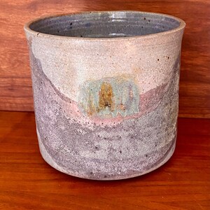May include: A ceramic planter with a light gray, purple, and brown glaze. The planter has a rounded shape and a slightly textured surface.