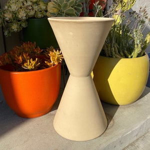 May include: A white ceramic hourglass-shaped planter with a wide opening at the top and a narrow base. The planter is sitting on a concrete surface with other potted plants in the background.