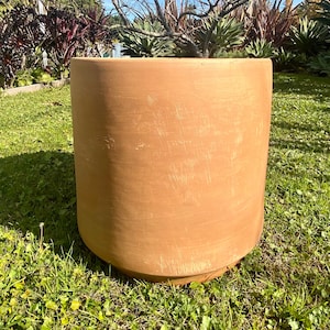May include: A large, terracotta-colored, cylindrical planter pot with a slightly rounded bottom. The pot is sitting on a bed of green grass.
