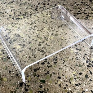 May include: A clear acrylic laptop stand with a curved front edge. The stand is designed to elevate a laptop or tablet for a more ergonomic viewing angle.