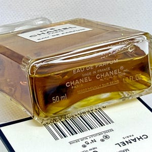 May include: A rectangular glass perfume bottle containing amber liquid, labeled Chanel No. 5 Eau de Parfum. The base shows 50 ml and 1.7 fl oz. The bottle rests on a white box featuring a barcode and the Chanel logo with text including Paris and New York.
