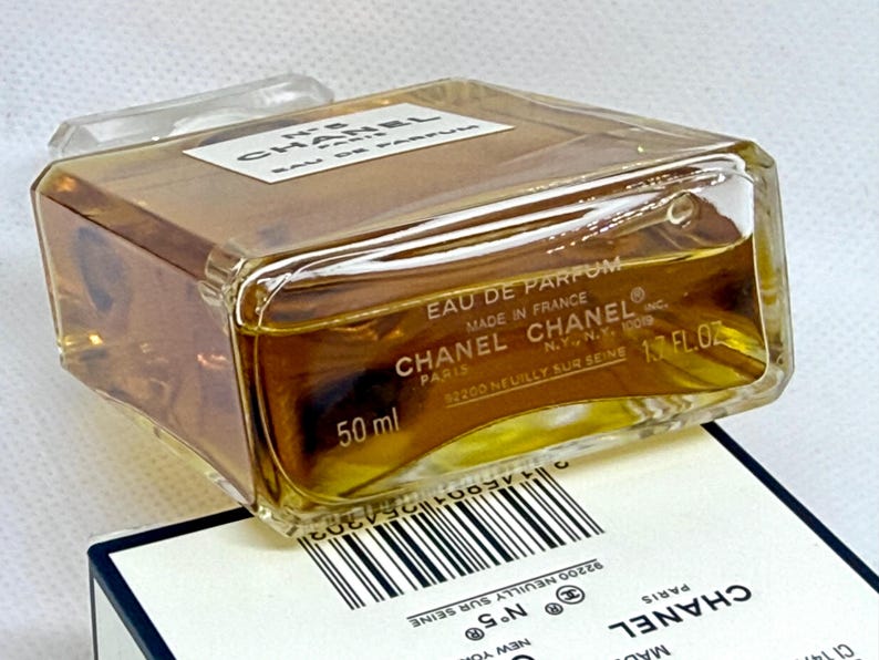 May include: A rectangular glass perfume bottle containing amber liquid, labeled Chanel No. 5 Eau de Parfum. The base shows 50 ml and 1.7 fl oz. The bottle rests on a white box featuring a barcode and the Chanel logo with text including Paris and New York.
