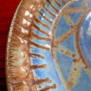 May include: Close-up of a ceramic plate with a brown rim and a blue and brown patterned center. The plate has a rustic, handmade appearance, with visible texture and variations in color.