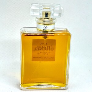 May include: A rectangular glass perfume bottle filled with amber-colored liquid. The label reads N&deg;5 CHANEL PARIS EAU DE PARFUM. It features a clear, faceted glass stopper and a gold-toned collar with a white band displaying the Chanel logo.
