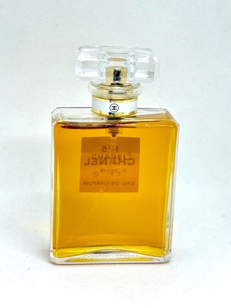 May include: A rectangular glass perfume bottle filled with amber-colored liquid. The label reads N&deg;5 CHANEL PARIS EAU DE PARFUM. It features a clear, faceted glass stopper and a gold-toned collar with a white band displaying the Chanel logo.